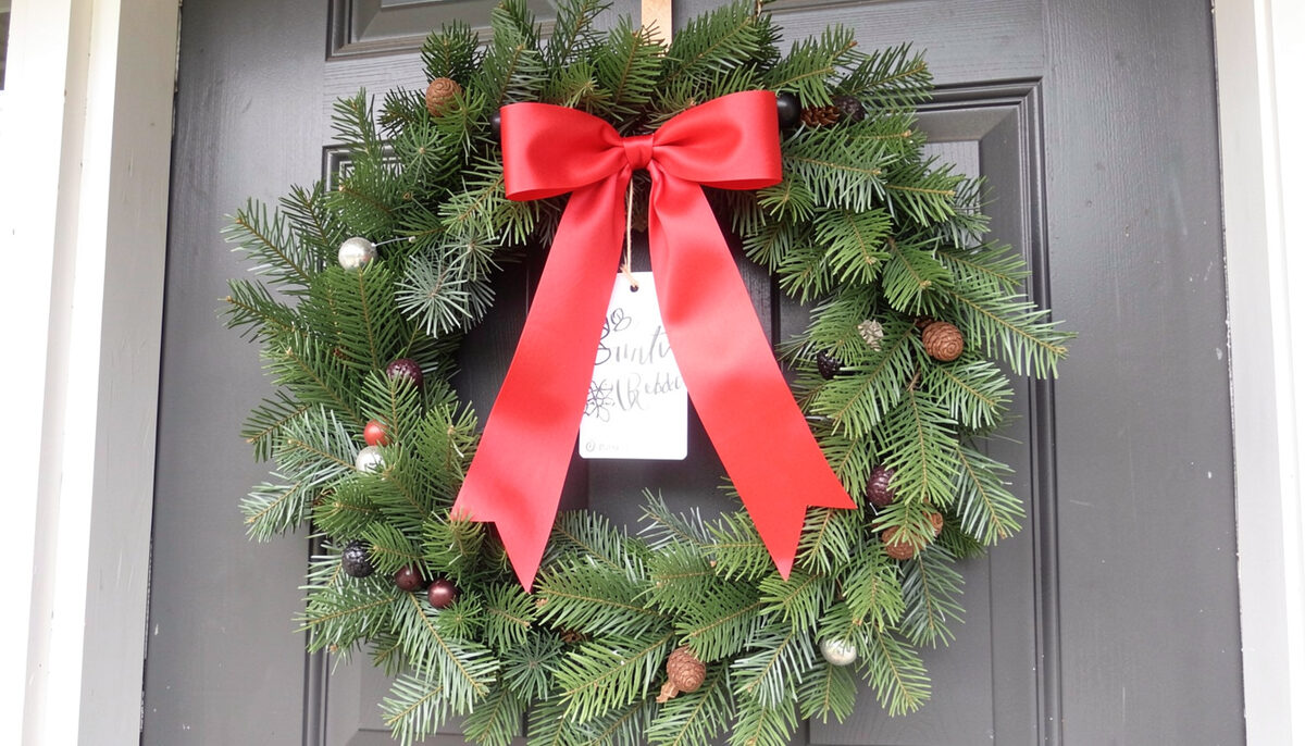 A fresh evergreen wreath hanging on a painted front door with a red bow, a paper gift tag tied to it, daylight.