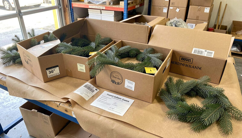 Several identical boxed evergreen wreaths on a shipping bench with kraft paper, address labels and a printed card, warehouse daylight.
