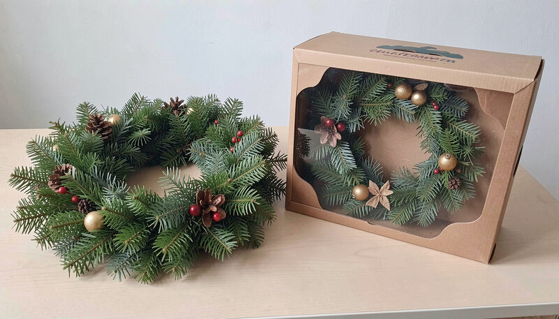 A fresh evergreen wreath and a boxed artificial wreath resting on a table beside each other, plain indoor light.