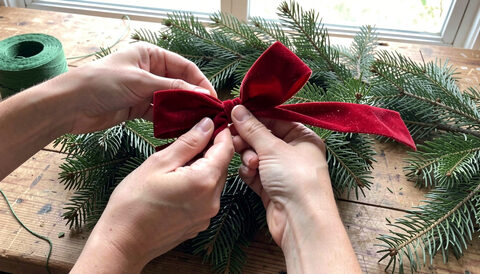 Two hands tying a red velvet bow onto a fresh noble fir wreath on a wooden workbench with pine needles and green florist wire nearby