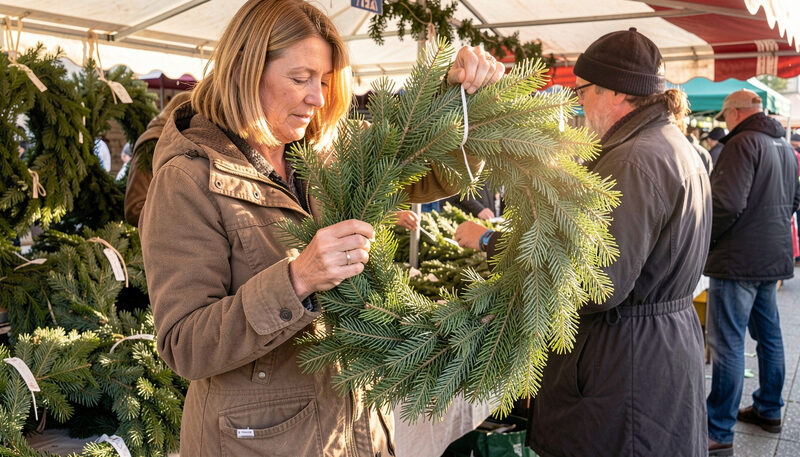 A person holding a fresh evergreen wreath up to the light at a market stall, checking the needles, with other wreaths stacked behind.